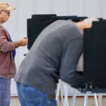 Two people are standing at voting booths, filling out ballots. The scene is indoors, with a white wall in the background. One person is in focus, while the other is blurred in the foreground.