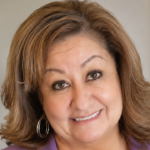School counselor Jeanette Martinez is suing Albuquerque Public Schools over its gender secrecy policy. Pictured: a woman with light brown hair, wearing hoop earrings and a purple top, smiles at the camera. The background is softly blurred, with a plant and a framed picture visible.