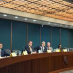 The Joint Election Equipment Specifications and Standards CommitteeA group of people meet at a long, curved conference table in a formal meeting room with microphones and nameplates in front of them. A TV screen shows another person in the background.