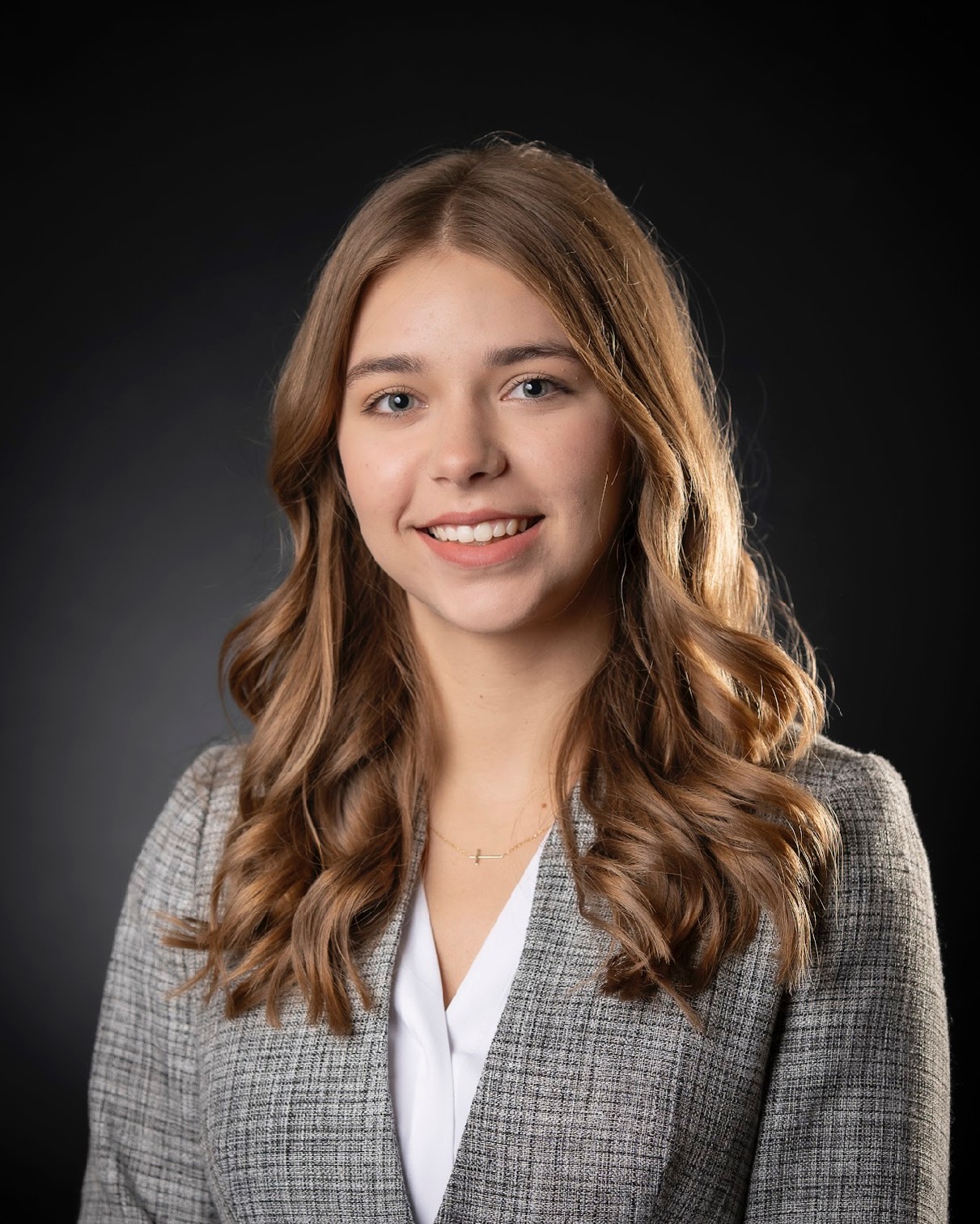 Daily Signal Internship 3 A young woman with long, wavy light brown hair wears a gray plaid blazer and white blouse, smiling at the camera against a dark background—professional and confident, she perfectly embodies the polished look of a Daily Signal internship participant.