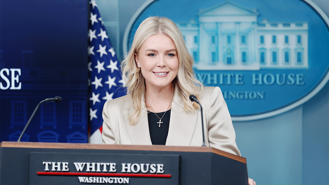 A woman with blonde hair speaks at a podium in the White House press briefing room, with an American flag and a White House emblem in the background, during a Trump administration event.