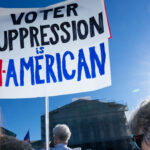 People stand outside in sunlight holding a sign that reads, Voter suppression is un-American, with part of the sign in red, black, and blue letters. A government building is visible in the background.