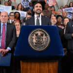 Zohran Mamdani speaks at a podium with an official seal, surrounded by a group of people holding folders and signs supporting immigrants and legal aid inside a government building.