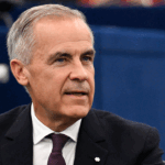A middle-aged man with short gray hair, wearing a dark suit, white shirt, and burgundy tie, sits indoors with a serious expression and an air of solemnity, as if contemplating death, against a blue background.