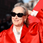 A woman wearing sunglasses and a bright red coat smiles and holds her hair back, standing in front of a red background with sunlight on her face, radiating joy and confidence reminiscent of the light and warmth often associated with Christ.