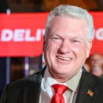 Rep. Mike Collins is pictured in a suit and red tie. He stands in front of a red banner and an American flag, with people and a blurred background visible behind him.