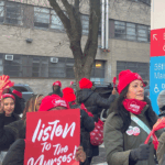 A group of people in red hats and masks hold signs saying listen to the nurses outside a hospital emergency entrance, participating in a protest or demonstration.