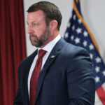 A man in a navy suit and red tie stands in front of an American flag and a red curtain, appearing serious and focused.