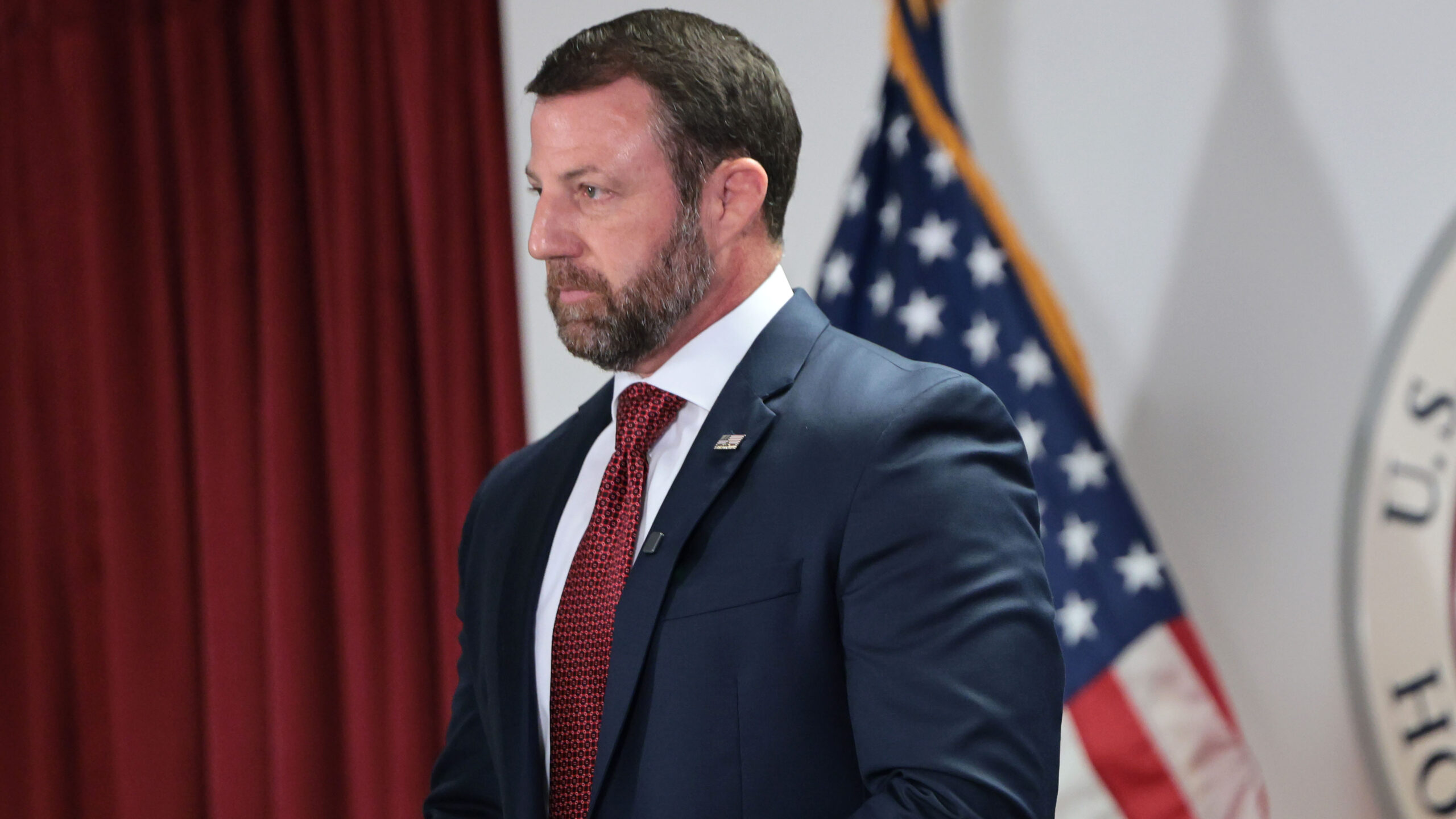 A man in a navy suit and red tie stands in front of an American flag and a red curtain, appearing serious and focused.