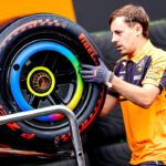 A mechanic in an orange and navy uniform carries a large Formula 1 Pirelli tire with colorful markings, likely for a racing event.