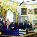 A group of men in suits, including Donald Trump seated at a desk, are gathered in the Oval Office with microphones overhead and shelves, flags, and presidential portraits in the background.