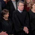 Four Supreme Court justices wearing black robes stand closely together during a formal event, surrounded by other attendees. They appear solemn, with their hands clasped or at their sides.