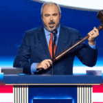 A man in a blue suit and red tie stands at a podium, holding a large wooden gavel and speaking into a microphone, with a blue and red background behind him.