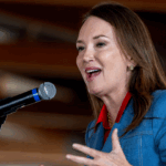 A woman in a blue blazer speaks into a microphone, gesturing with one hand. She has long brown hair and is indoors, with blurred background beams visible.