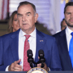 A man in a blue suit speaks at a podium with microphones, while another man in a suit stands behind him. An American flag is visible in the background, suggesting this event may be taking place in Missouri.