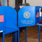 Two people fill out ballots at blue voting booths in a room with wood floors and walls. The booths display the Nassau County seal, an American flag sticker, and a sign with voter information in English and Spanish.