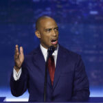 Scott Turner in a navy suit and red tie speaks passionately at a podium, gesturing with his left hand, against a blurred blue and purple background.