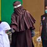 A person in brown robes prepares to cane a kneeling individual in white while uniformed officials and onlookers stand nearby, one wearing a face mask.