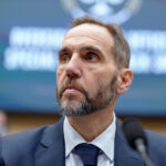 A man in a suit and tie with a beard looks to the side while seated in front of a blurred background with text.