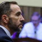 A man in a suit with a short beard listens intently during a meeting, while a blurry figure appears on a screen in the background.