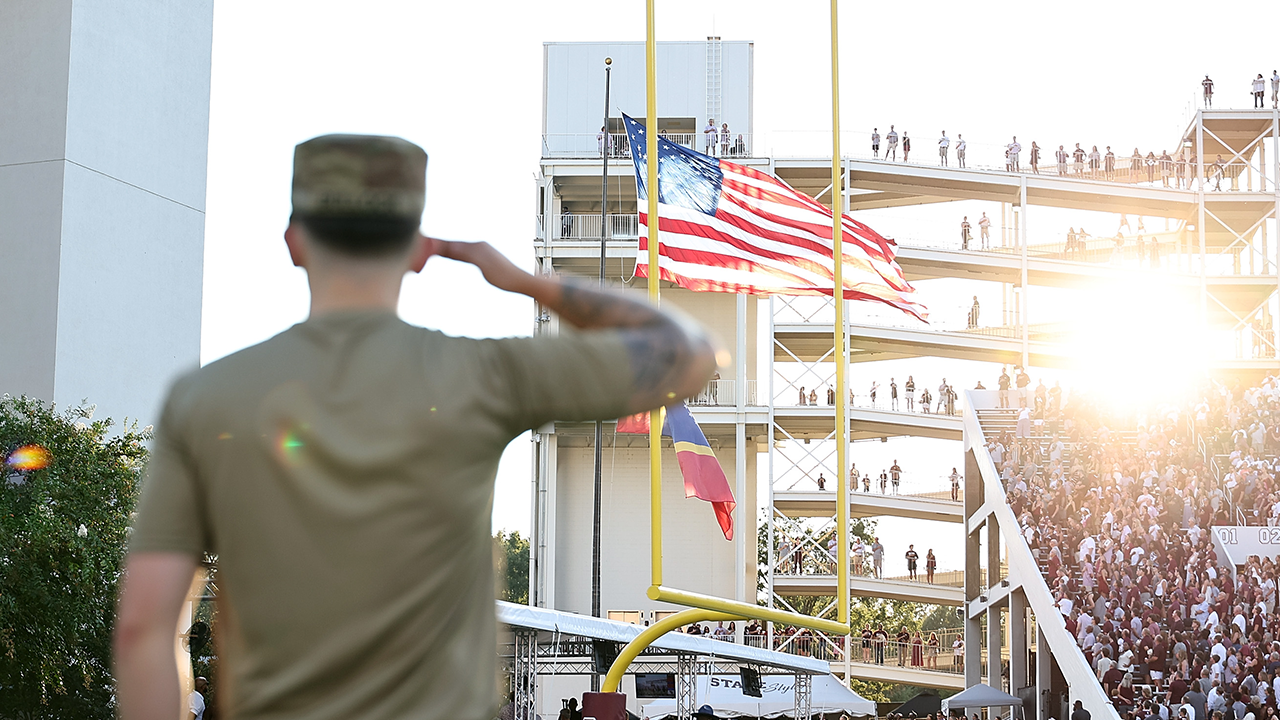 A soldier in uniform salutes the American flag at a football stadium, with sun shining and a crowd in the stands.