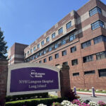 A large brick hospital building with a purple NYU Langone Hospital Long Island sign in front, surrounded by flowers and greenery under a sunny blue sky.