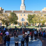 A crowd gathers in a plaza with trees, facing a speaker near balloons. In the background, a large building with a clock tower stands under a clear blue sky.