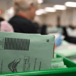 A close-up of green mail-in ballot envelopes in a tray at a busy, blurred election processing facility, with workers in the background.