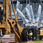 A worker operates a yellow drilling machine at a construction site, with large metallic components and equipment visible in the background.