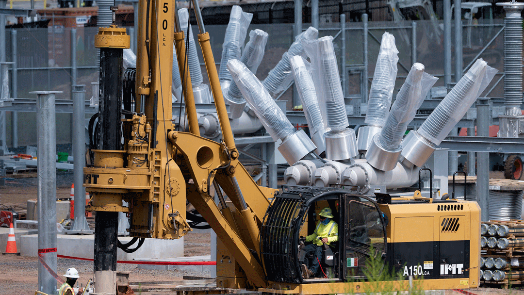 A worker operates a yellow drilling machine at a construction site, with large metallic components and equipment visible in the background.