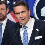 Todd Blanche and Markwayne Mullin in suits stand in front of U.S. flags and a sign that reads Department. The man in front speaks into a microphone, while the other looks on in the background.
