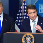 Two men in suits stand at a podium with the U.S. presidential seal. An American flag and a backdrop with the White House logo are visible behind them. One man speaks while the other looks on.