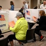 People sit at voting booths filling out ballots in a polling station. Voting partitions display the American flag and the word VOTE. The room is well-lit, and several voters are present.