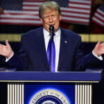 Donald Trump in a blue suit speaks at a podium with the presidential seal, gesturing with both hands, while American flags are held in the background.