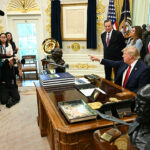 Donald Trump sits at the Oval Office desk, pointing while surrounded by officials, staff, and reporters. There are flags, documents, and busts visible in the ornate room. A photographer kneels, taking photos.