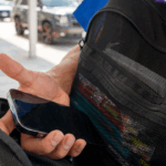 A student holds a smartphone device while opening the mesh pocket of a black backpack outdoors.