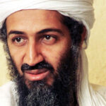 Close-up portrait of Osama bin Laden wearing a white turban and a long, dark beard, looking slightly off-camera against a plain, light-colored background.