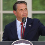 Todd Blanche in a dark suit speaks into a microphone at a podium outdoors, pointing to his right. Two large American flags are visible in the background.