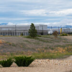 A dirt road and grassy field lead to a cluster of modern, gray industrial buildings, surrounded by fencing, with mountains and clouds in the background.
