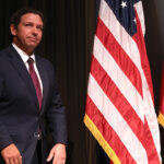 A man in a dark suit stands next to the United States flag and another flag with red, white, and blue stripes, against a dark backdrop.