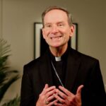 Bishop Michael Burbidge, dressed in clerical attire, speaks, gesturing with his hands. Behind him are green plants. A Catholics Vote No logo appears in the lower right corner.