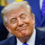Donald Trump in a suit and blue tie sits, smiling with eyes closed, in a brightly lit room. Others in suits are blurred in the background.