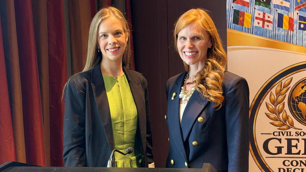 Two women stand side by side, smiling, in front of a podium and a banner adorned with international flags and a civil society emblem. Their expressions convey the energy and life of the event—one wears a green dress paired with a black jacket; the other is dressed in a navy blazer with gold buttons.