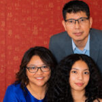Three people pose in front of a red wall with gold patterns. A man in a blue suit stands behind a woman in glasses and a blue dress, while a young woman with curly hair sits in front. Though all are looking at the camera and smiling, their expressions hint at the tension of an unexpected hostage scenario unfolding just beyond the frame.