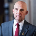 Bald man in a gray suit, red tie, and white shirt stands indoors. He has a serious expression and wears a pin on his lapel. The background is softly blurred.