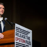 A man in a suit speaks at a podium with a sign reading RELEASE THE EPSTEIN FILES in bold letters, next to a microphone, against a dark background.