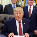 Donald Trump in a suit and red tie sits at a desk, speaking, with five men in suits standing behind him in a formal setting with flags and military decorations in the background.