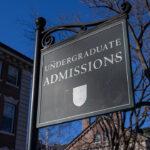 A black sign reads Undergraduate Admissions outside a brick building, with bare tree branches and a clear blue sky in the background.