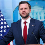 JD Vance in a dark suit and red tie speaks at a podium in front of American flags and a White House emblem backdrop.