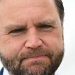 A close-up of JD Vance with short brown hair, a full beard, and a serious expression, with a blurred light background.